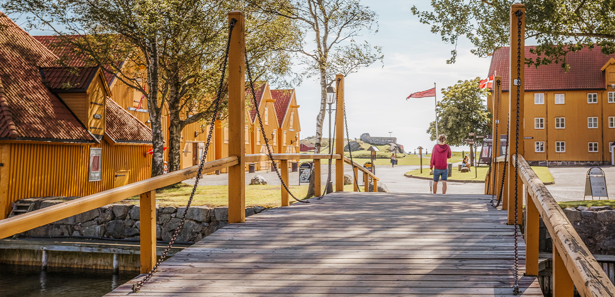 Trebro i Stavern med gule trehus i bakgrunnen. Broen er laget av planker med gule rekkverk og kjettinger. Tradisjonelle gule bygninger med røde tak ligger langs vannet, og en person i rød genser går i det fjerne.