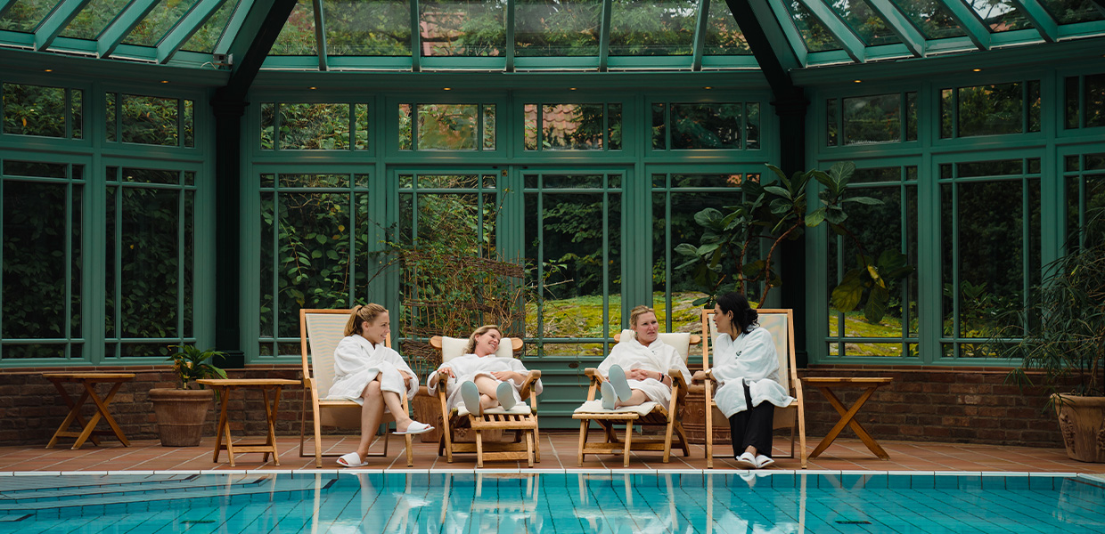 fire damer lener seg tilbake på solstoler i et art deco- stil innendørs bassengområde. Grønne vegger med store vinduer rammer dem inn, det gir en slags drivhus-effekt. Du skimter bassenget i forgrunnen og grønn vegetasjon i bakgrunnen.