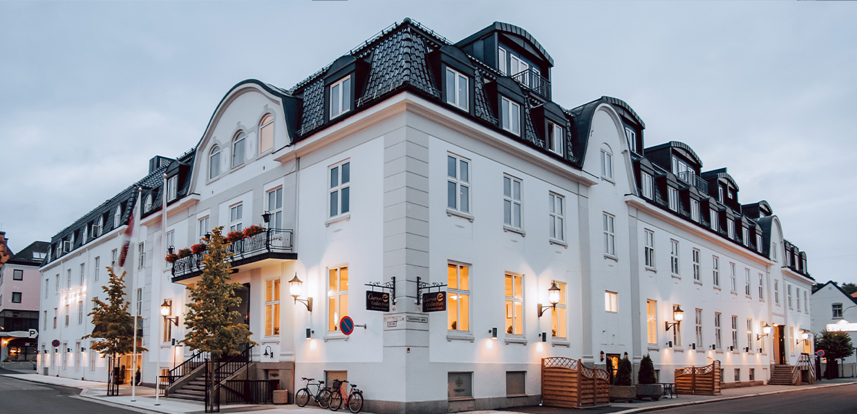 Stilfull bygning i hvitt, med mørkt tak og store vinduer. Kveldsmørket har senket seg, og fasaden er opplyst av koselige utelamper.