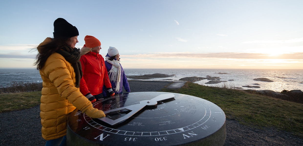 Tre kvinner i fargerike vinterjakker står ved et kompasskilt med havet i bakgrunnen.