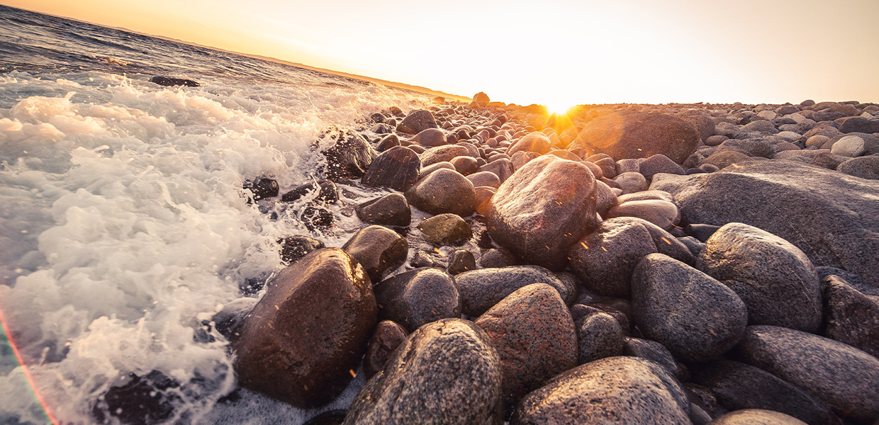 En steinete strandlinje ved solnedgang, hvor bølgene fra havet skvulper opp mot de glatte, runde steinene som ligger tett i tett langs kysten. Solen er i ferd med å gå ned i horisonten, og kaster et gyllent lys over landskapet, noe som får steinene til å skinne i varme toner av oransje og gull. Havet er lett brusende, og man kan se små bølgeskum nærmest i bilde