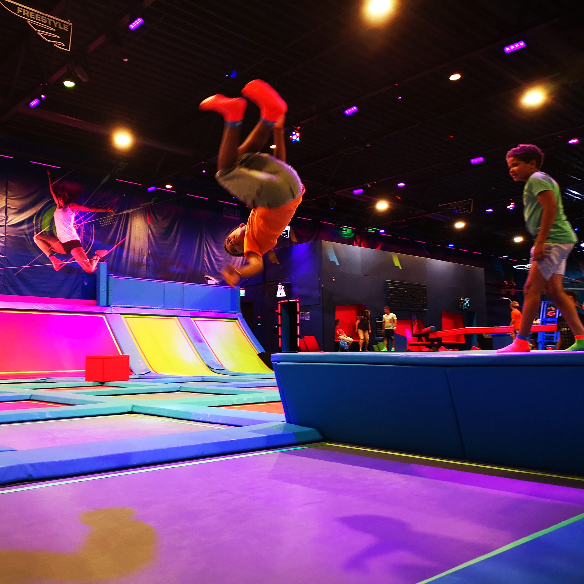 Ungdommer utfører akrobatiske hopp på trampoliner i en fargerik lyssetting.