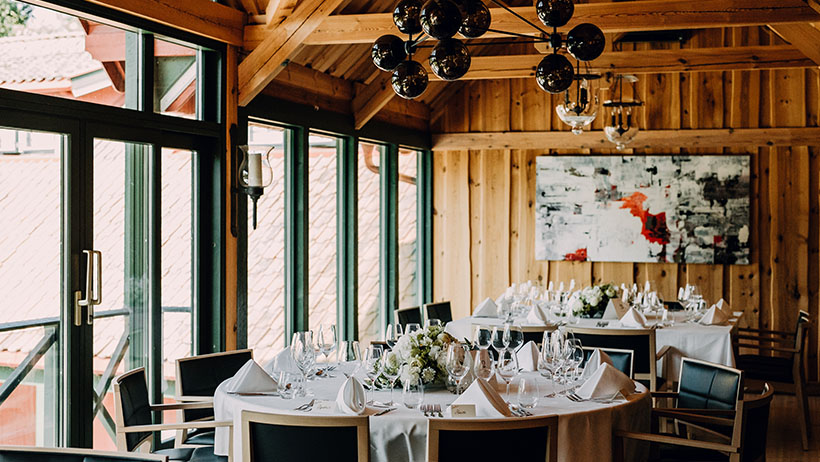 Et selskapslokale med runde bord dekket med hvite duker, vinglass og blomsterdekorasjoner. En stor lampe henger fra tretaket, og vinduene slipper inn dagslys og gir utsikt til bygninger av rødt treverk utenfor.