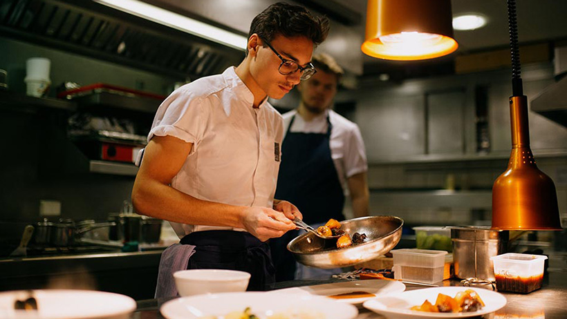 To kokker står på et restaurantkjøkken og legger mat fra en stekepanne over på tallerkener.