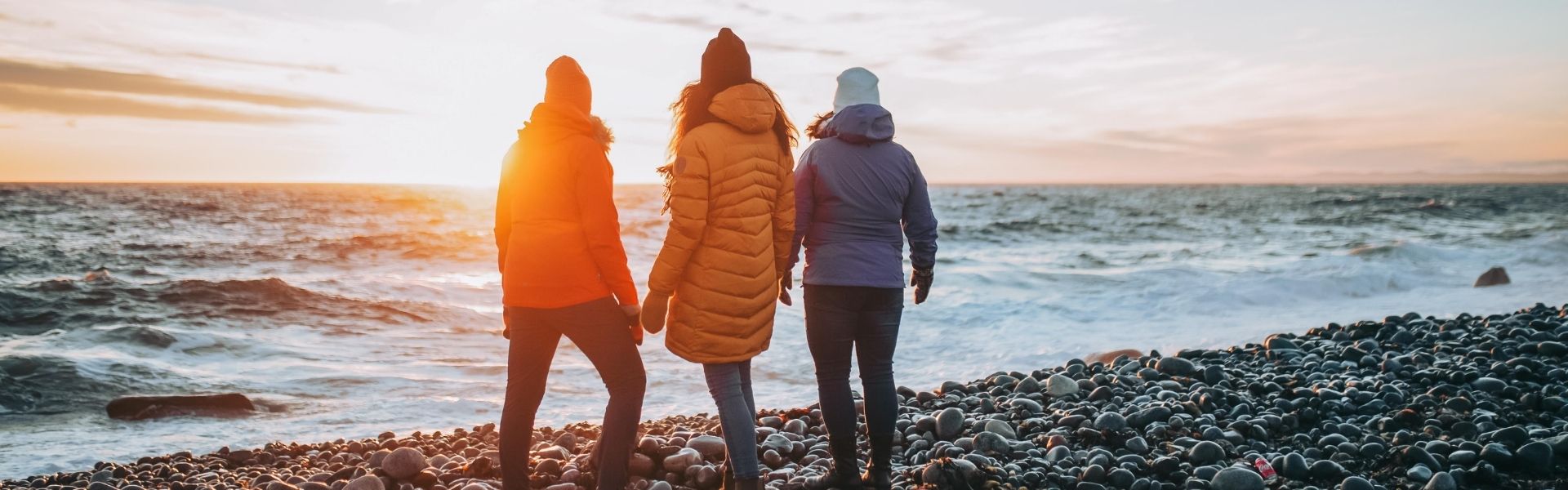 Tre personer ser på solnedgang ved havet stående på rullesteiner