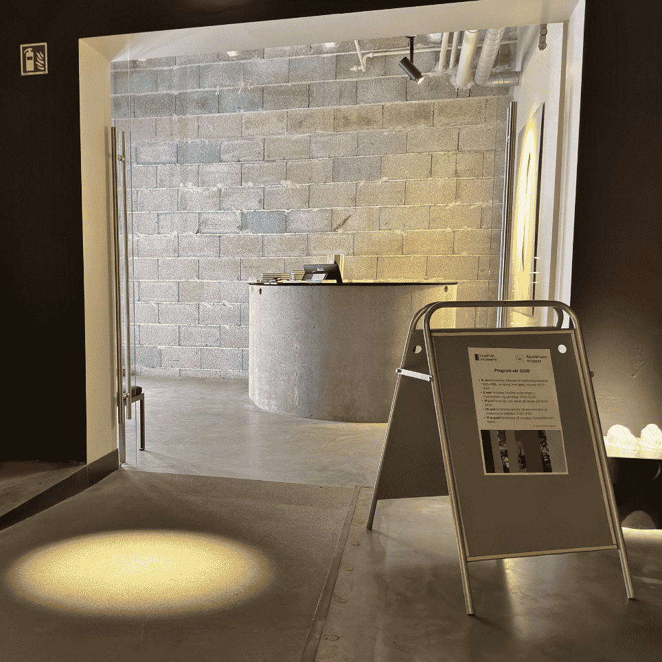 Entrance area with a glass door leading into a foyer featuring a grey concrete wall, a round counter and a poster stand, with warm lighting on the floor.