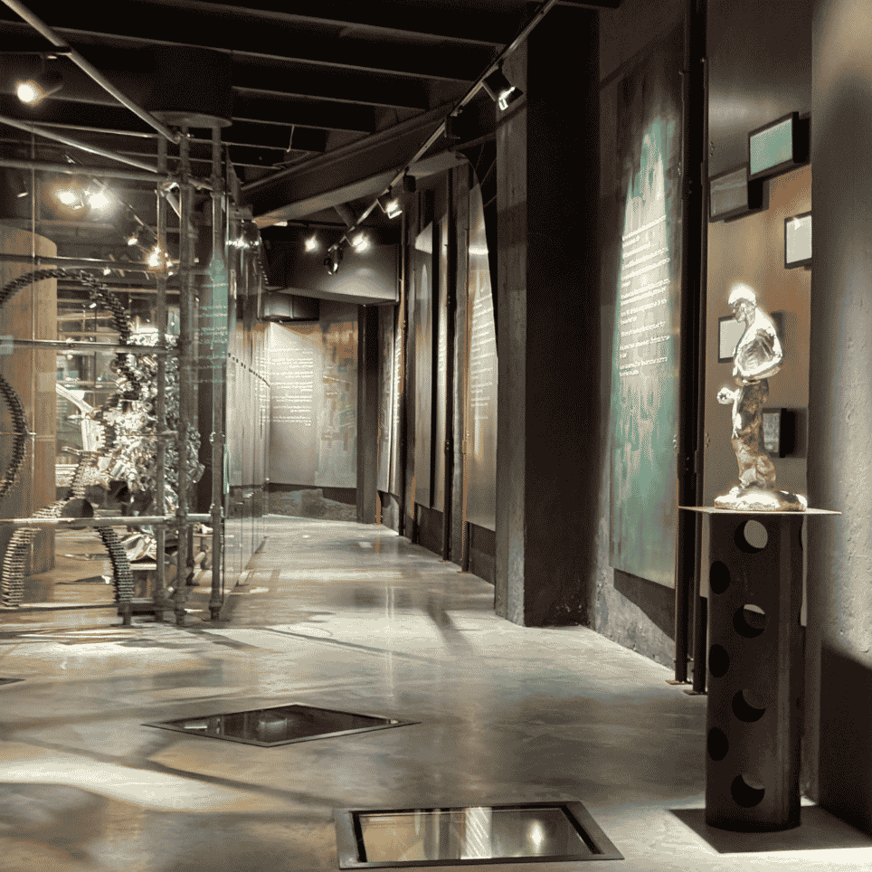 A dark exhibition space with metal sculptures behind glass, illuminated text posters on the wall and a statue on a plinth.