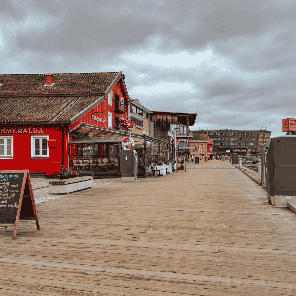 Trebrygge langs rødmalte sjøhus, med en tavle med meny og uteservering.