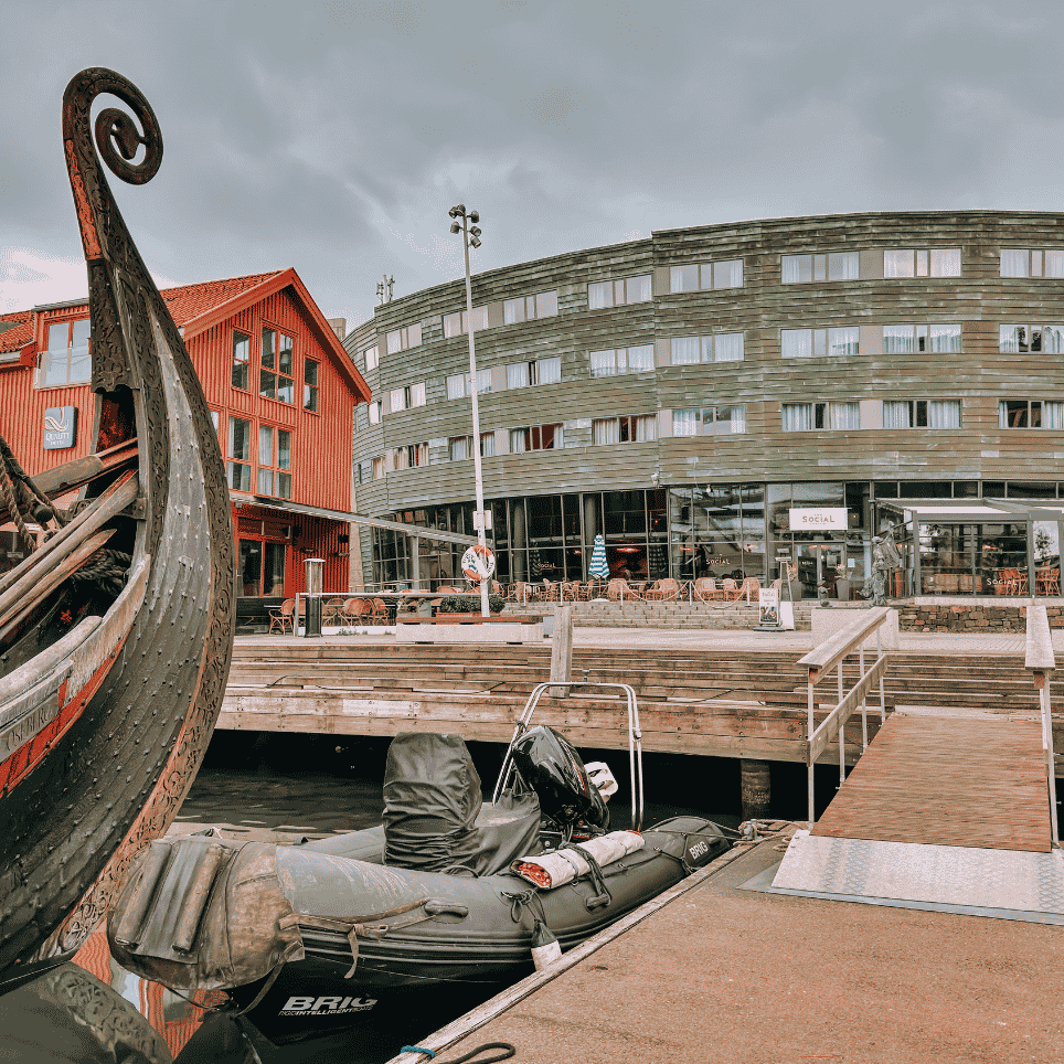 Nærbilde av baugen på et gammelt vikingskip, med en rødmalt bygning og en rundet moderne hotellbygning i bakgrunnen.