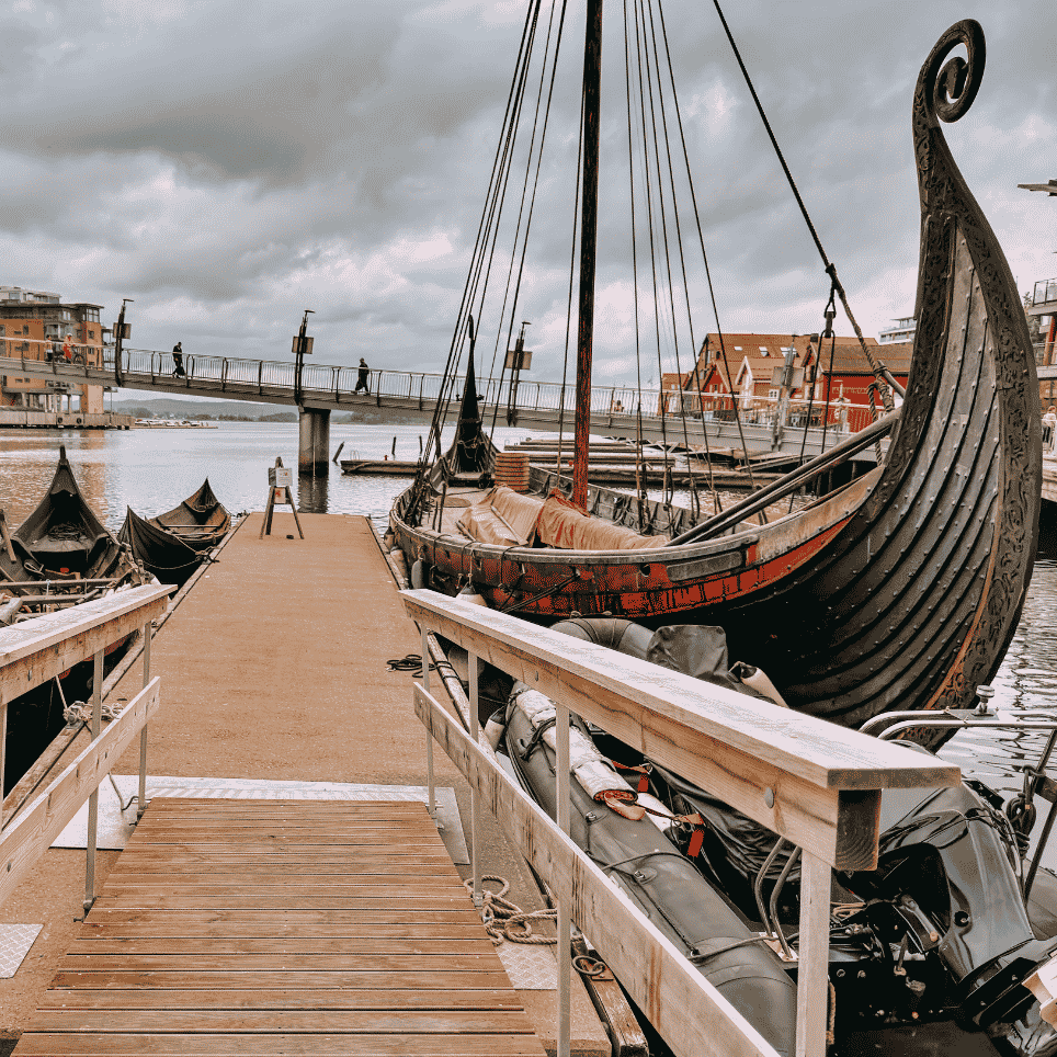 Gammelt seilskip fortøyd ved brygge, med mindre båter, en gangbro og boligblokker i bakgrunnen.