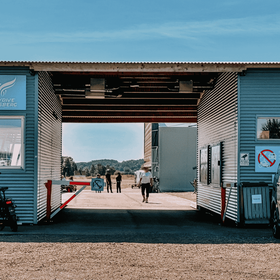 Inngangsparti til en blå hangarbygning med åpen port, bom og personer som går mot en flyplass, med en bil parkert utenfor.