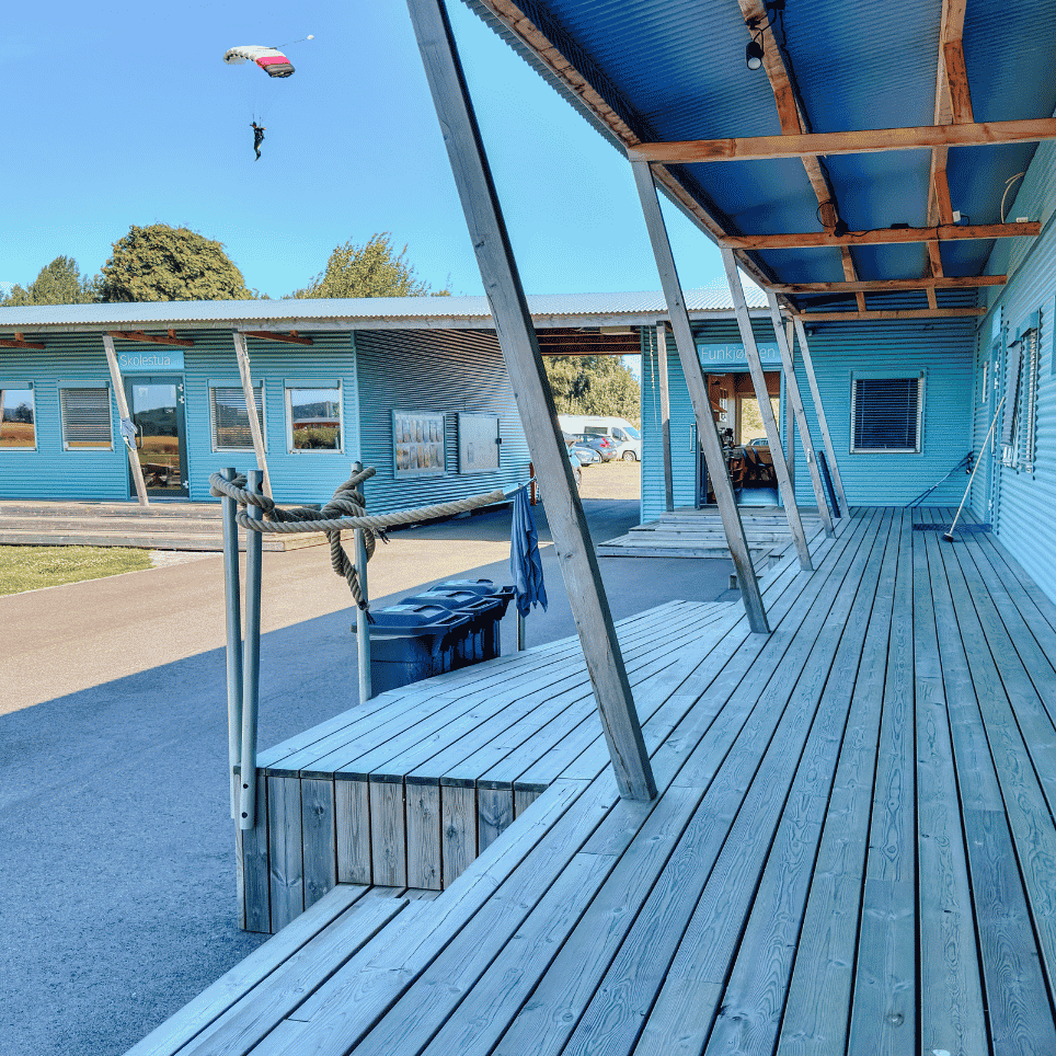 Trerekkverk og terrasse utenfor en blå bygning, med en fallskjerm synlig langt oppe på himmelen.
