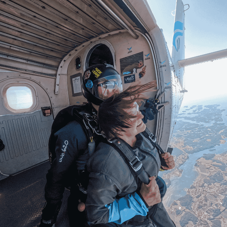 To personer klare til å hoppe ut av et fly, med utsikt over kyst og landskap under.
