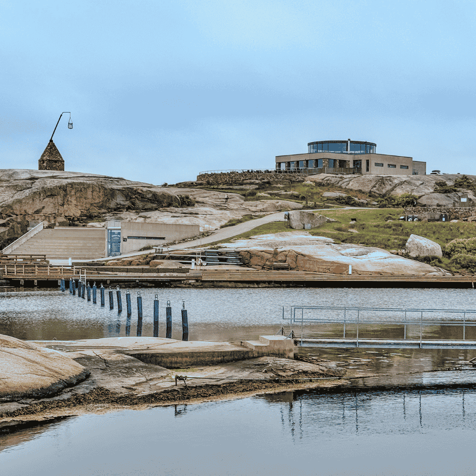 Vid utsikt over svaberg med en steinhytte og et moderne besøkssenter øverst, med brygge og molo nedenfor.