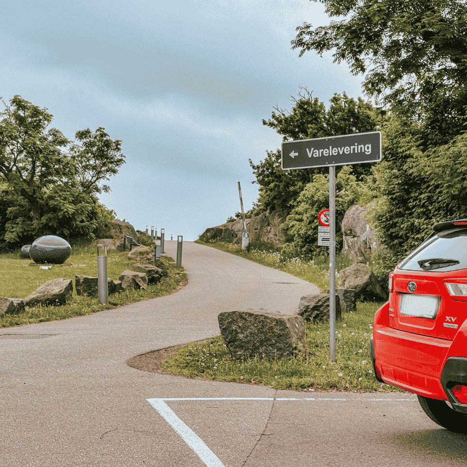 Rød bil parkert ved et skilt for varelevering, med en rund steinskulptur og en gruset sti i bakgrunnen.