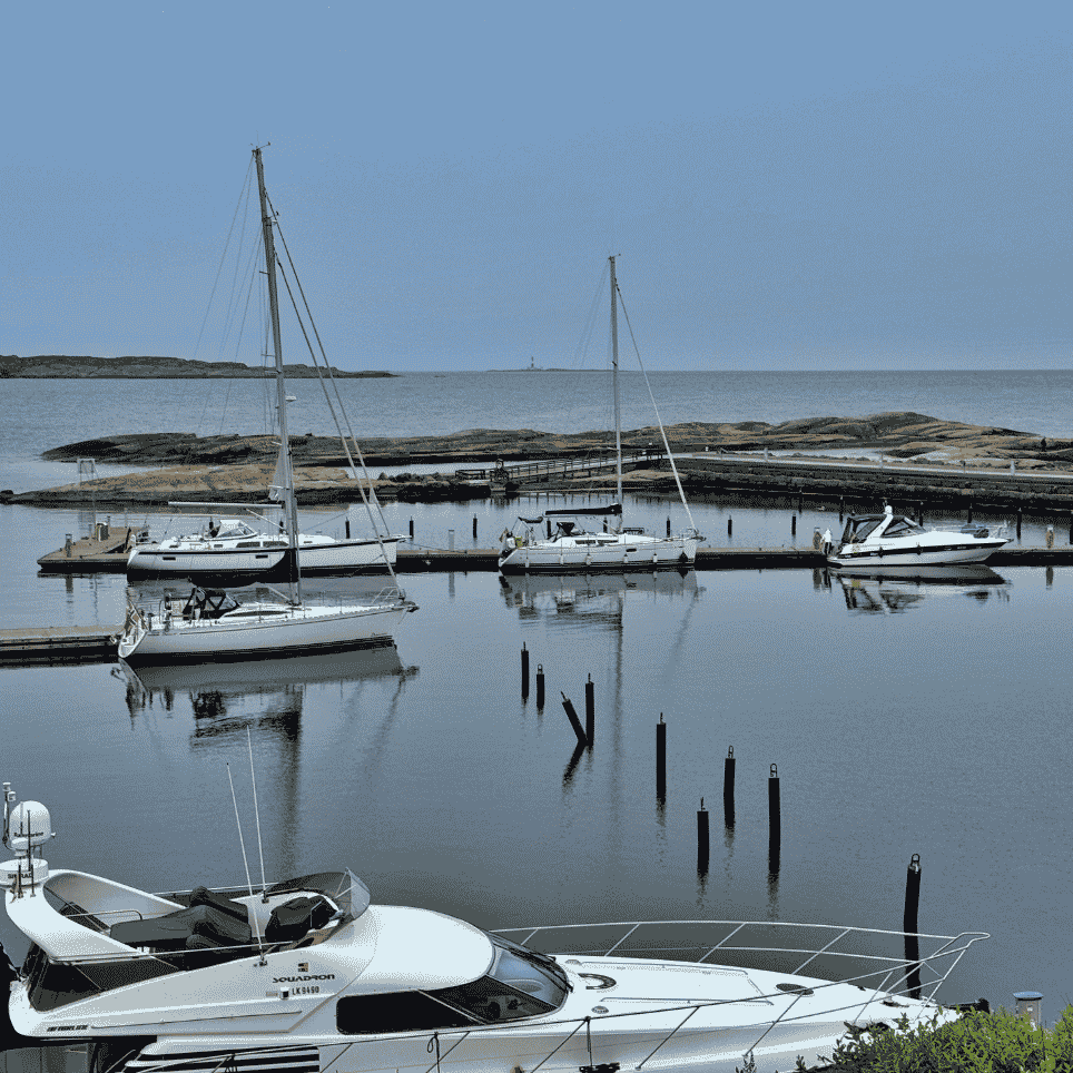 Småbåthavn med seilbåter fortøyd ved brygger, en motorbåt i forgrunnen og et fyr synlig langt ute ved horisonten.