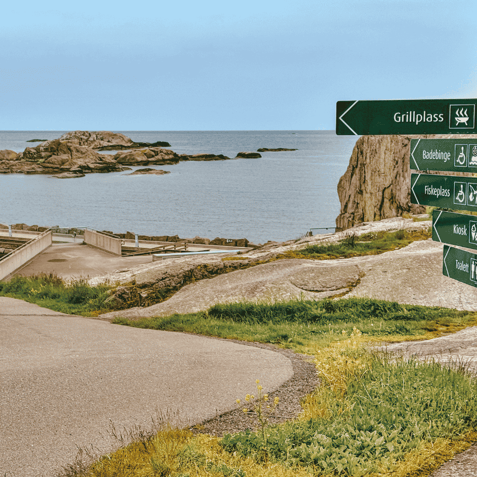 Skiltstolpe med retningsskilt til grillplass, badebinge, fiskeplass, kiosk og toalett, med havet i bakgrunnen.