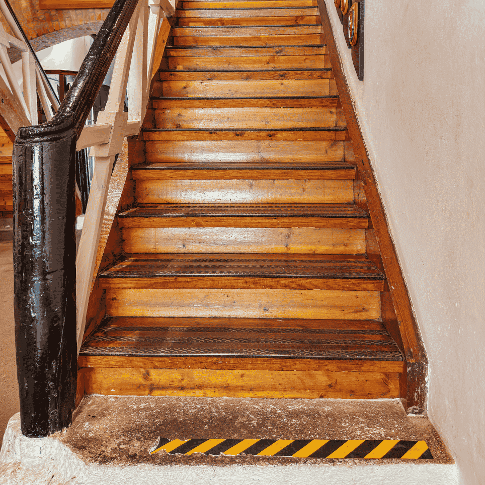 A steep wooden staircase with a black banister and fluted edge fittings, featuring a yellow-and-black warning stripe at the foot of the stairs.