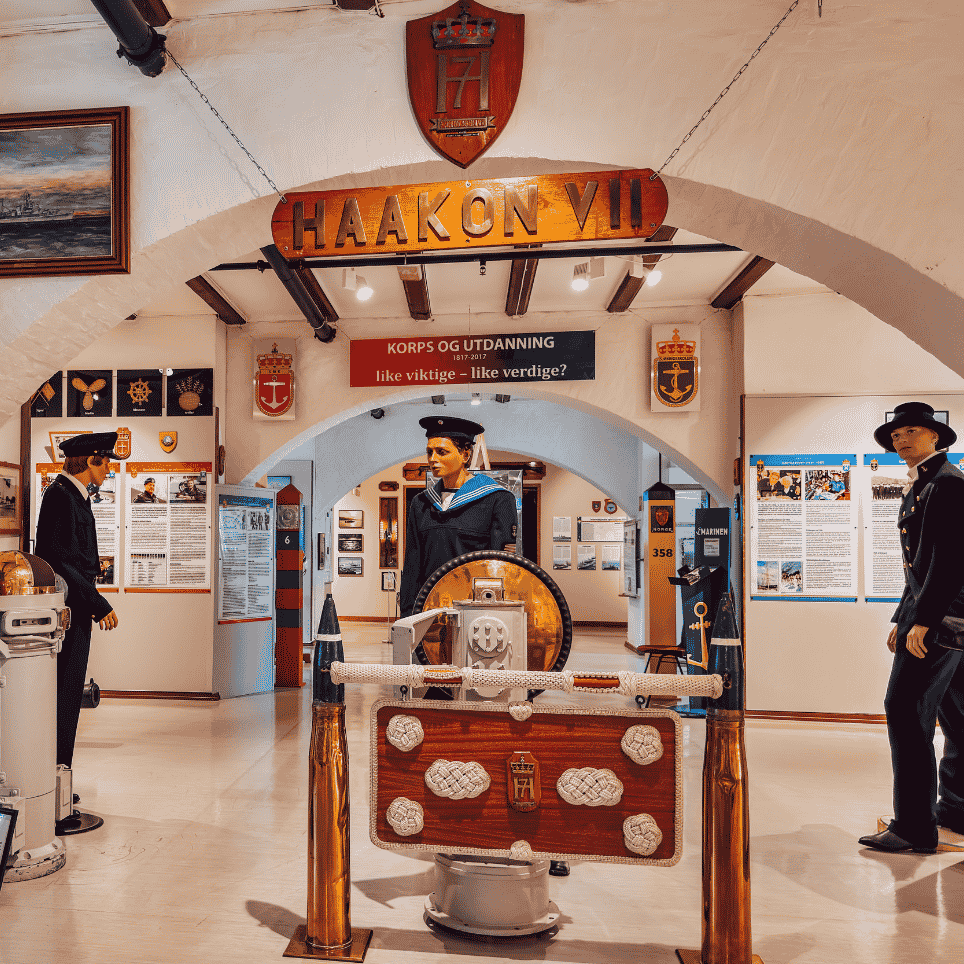 A museum room with mannequins in naval uniforms beneath a suspended arched sign, with information posters and coats of arms on the walls.