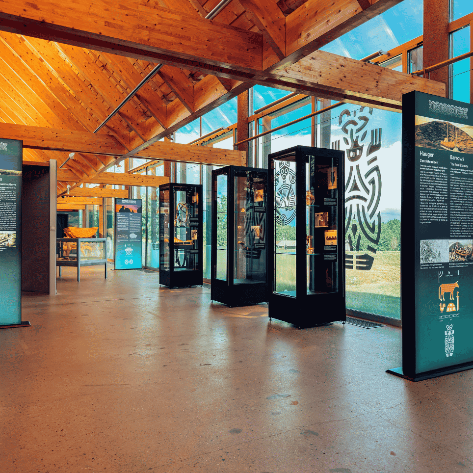 A museum shop with a sloping beamed ceiling, glass display cases containing jewellery and souvenirs, and shields on a display wall by the entrance.