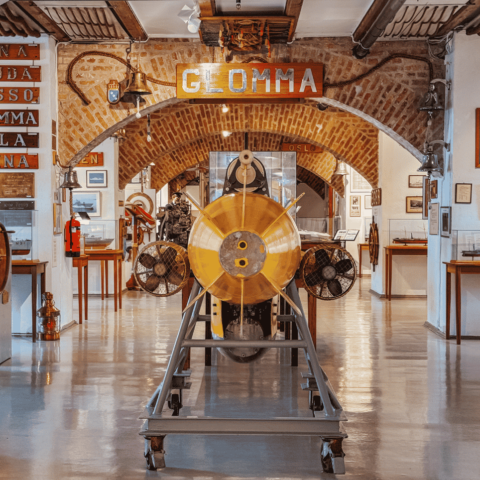 An exhibition room with a brick vaulted ceiling, featuring a yellow torpedo on wheels and ship’s nameplates hanging from the ceiling.