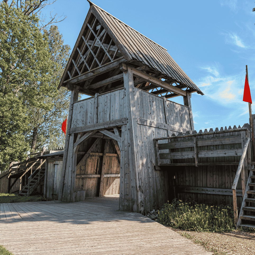 A three-storey watchtower with a pointed roof and two external staircases, forming part of a palisade with red flags and a gateway at the front.