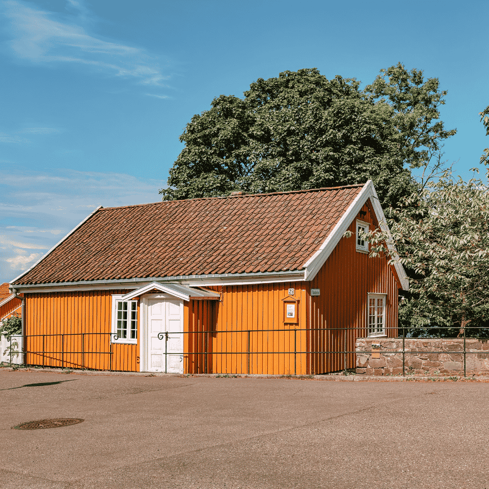 A yellow timber-panelled building with a tiled roof, a white front door and a free-standing tree in front of it, with a gravel surface and a manhole cover in the foreground.