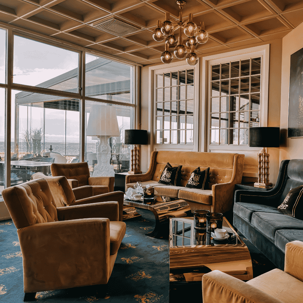A lounge with gold-coloured and dark velvet furniture, wall art and a chandelier, overlooking the sea.