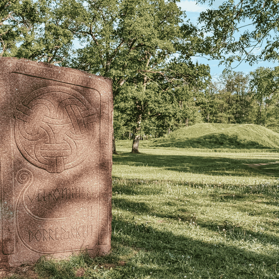 A standing stone with carved ornamentation, with a burial mound visible in the background.
