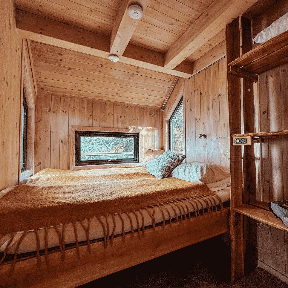 A loft bed with a yellow woollen blanket, windows on both sides and a shelf with bags next to it.