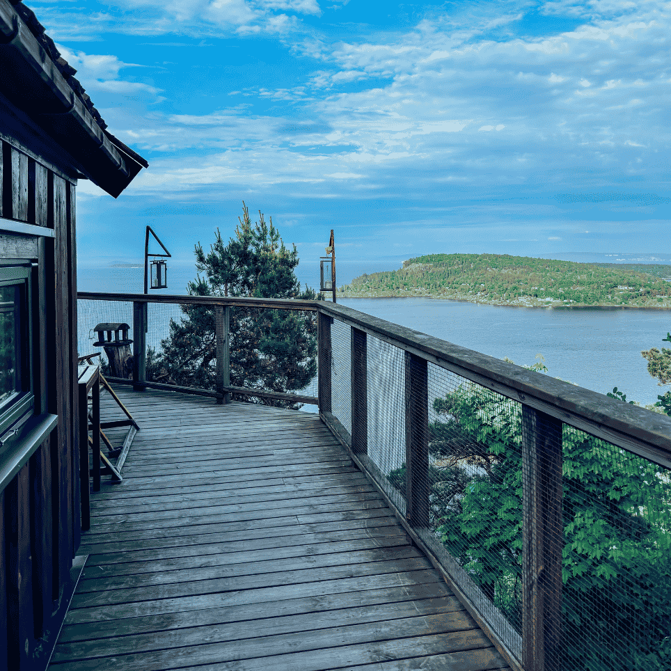 A terrace with a wooden chair, lanterns on the railing and a view of the water and pine trees.