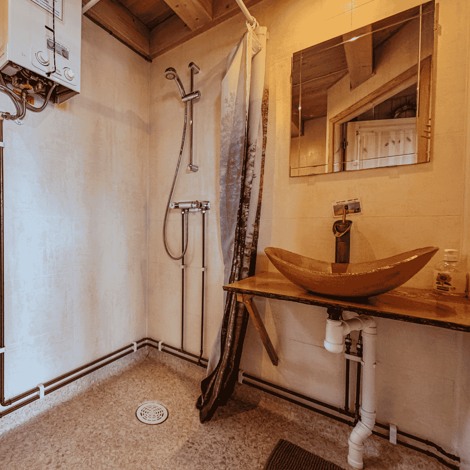 A bathroom with exposed pipework, a shower and an oblong wooden washbasin, with a mirror on the wall.