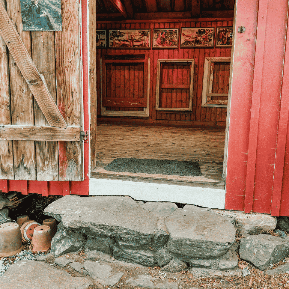 An open door leading into a red-painted gallery room with picture frames on the walls and a rug inside a stone threshold, with terracotta pots placed outside.