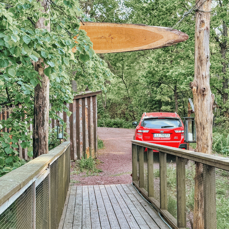 A wooden bridge with a carved sign above the road, and a red car parked a little further on amongst the trees.