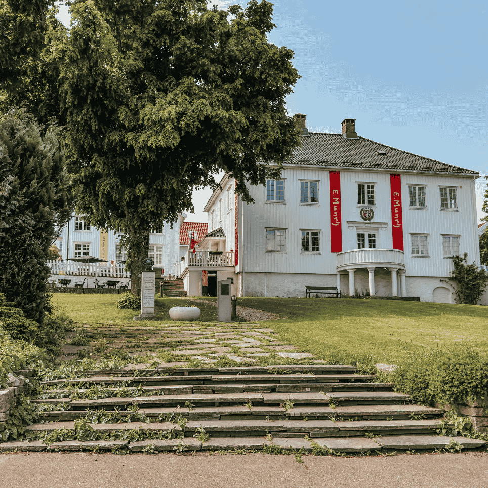 A white-painted building with red banners on the façade, seen from a stone staircase with a lawn and trees in the foreground.