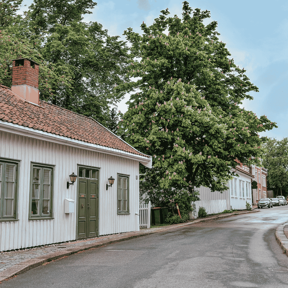 Hvitt trehus med rødt tegltak ved en svingete gate med et stort kastanjetre.