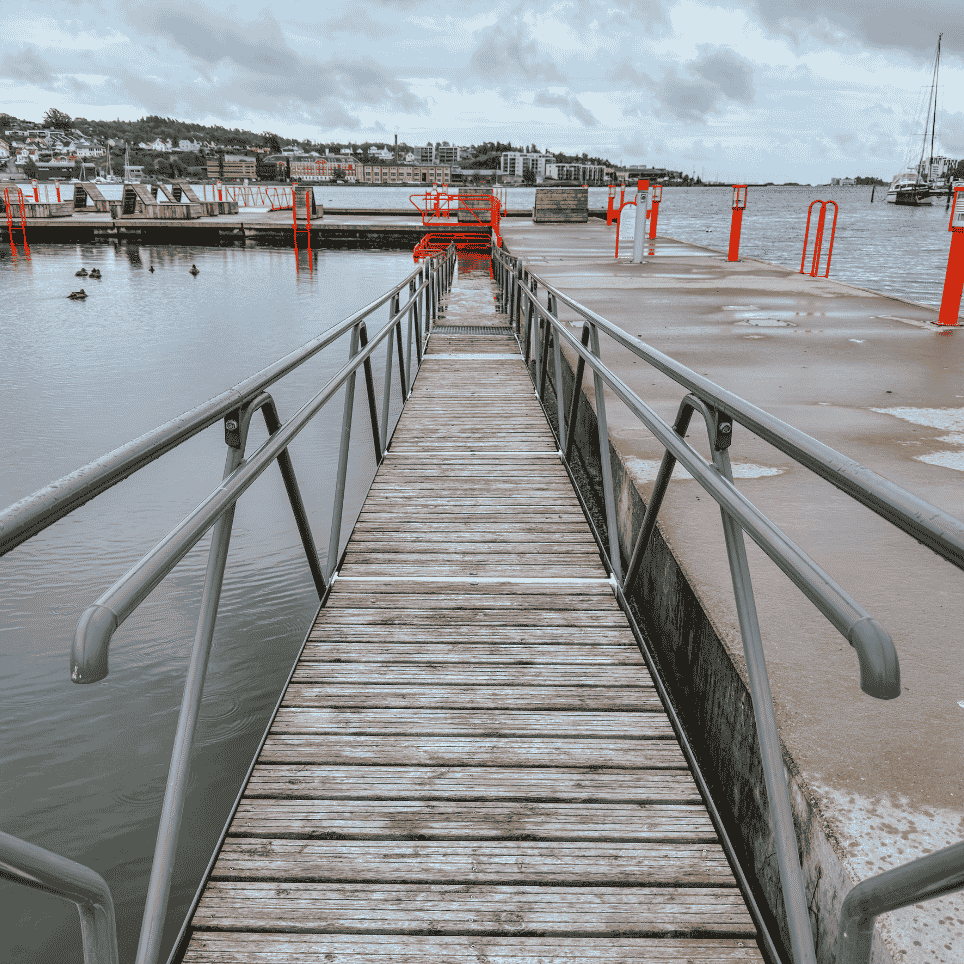 Trebrygge med rekkverk som leder ut til flytende badeplattformer med oransje sikkerhetsutstyr i havnen.
