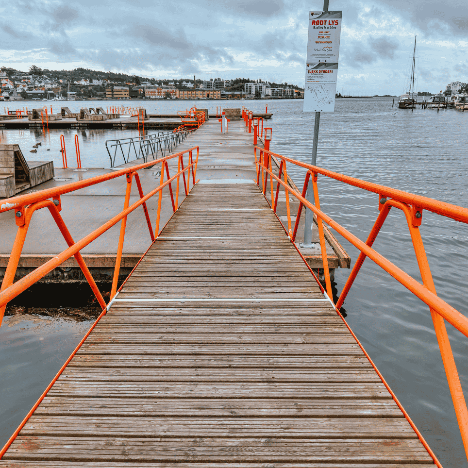 Brygge med oransje rekkverk og et skilt som varsler "RØDT LYS – Bading frarådes" ved badeanlegget.