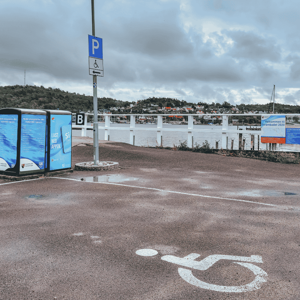 Handikapparkeringsplass ved havnen med en hurtigbåt merket "HYBRID" og informasjonstavler i bakgrunnen.