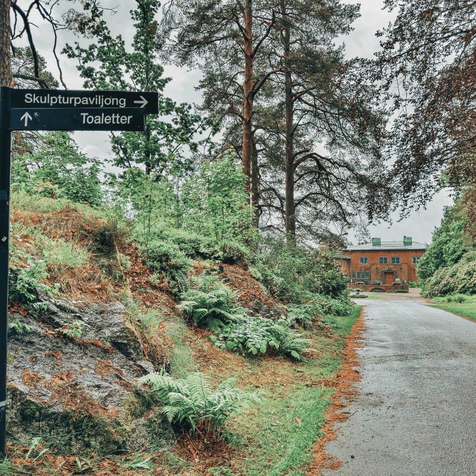 Skilt som peker mot "Skulpturpaviljong" og "Toaletter" ved en bregnebevokst skråning, med den rødmalte herskapsvillaen synlig for enden av gangveien.
