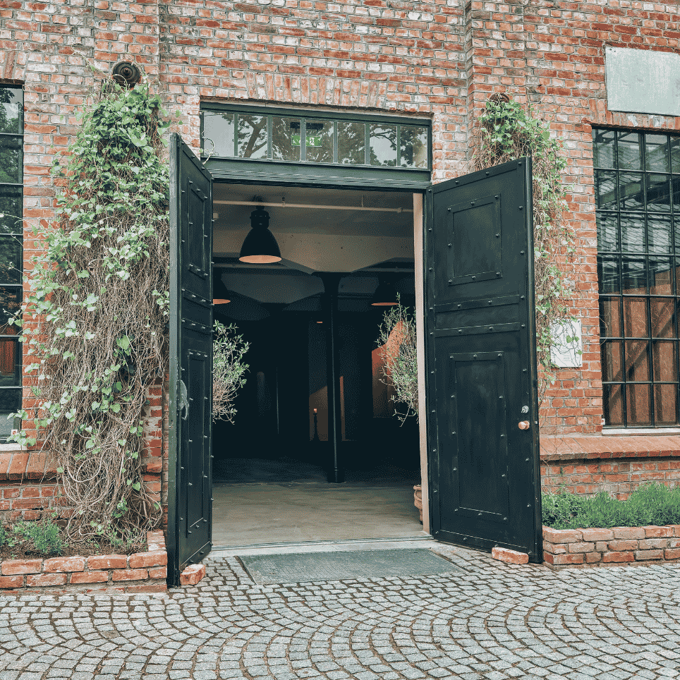 An open, black-painted double door leading into a brick building, with climbing plants along the wall and cobblestones in front of the entrance.