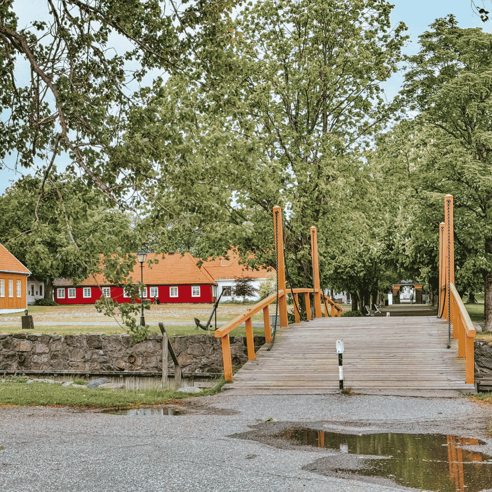 A yellow and red wooden building next to a fortress, with a yellow wooden bridge with a chain railing leading across a moat.