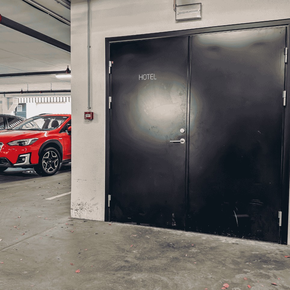 An underground car park with a black double door marked ‘HOTEL’, and a red and a black car parked in the background.