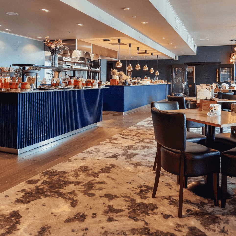 A restaurant area with a dark blue bar counter, gold-coloured pendant lights and tables with black upholstered chairs on a patterned carpet.