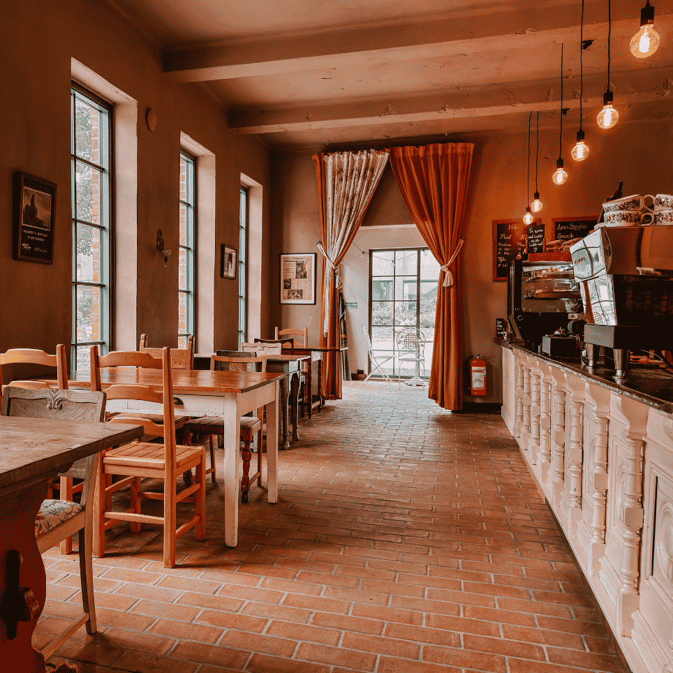A rustic café with a brick floor, wooden tables and chairs, orange curtains and a coffee bar with hanging light bulbs.