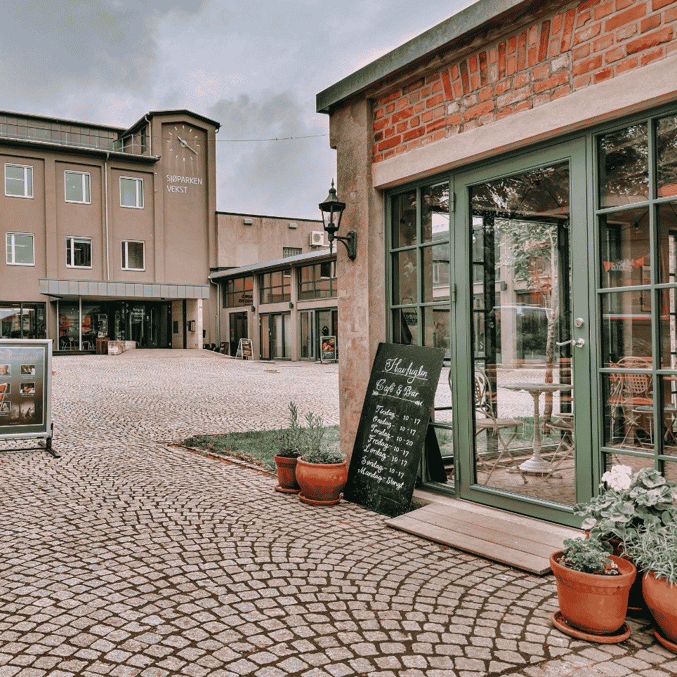 A paved courtyard with café tables, a sign for ‘Havfuglen café & bar’ and the ‘Sjøparken vekst’ building in the background.