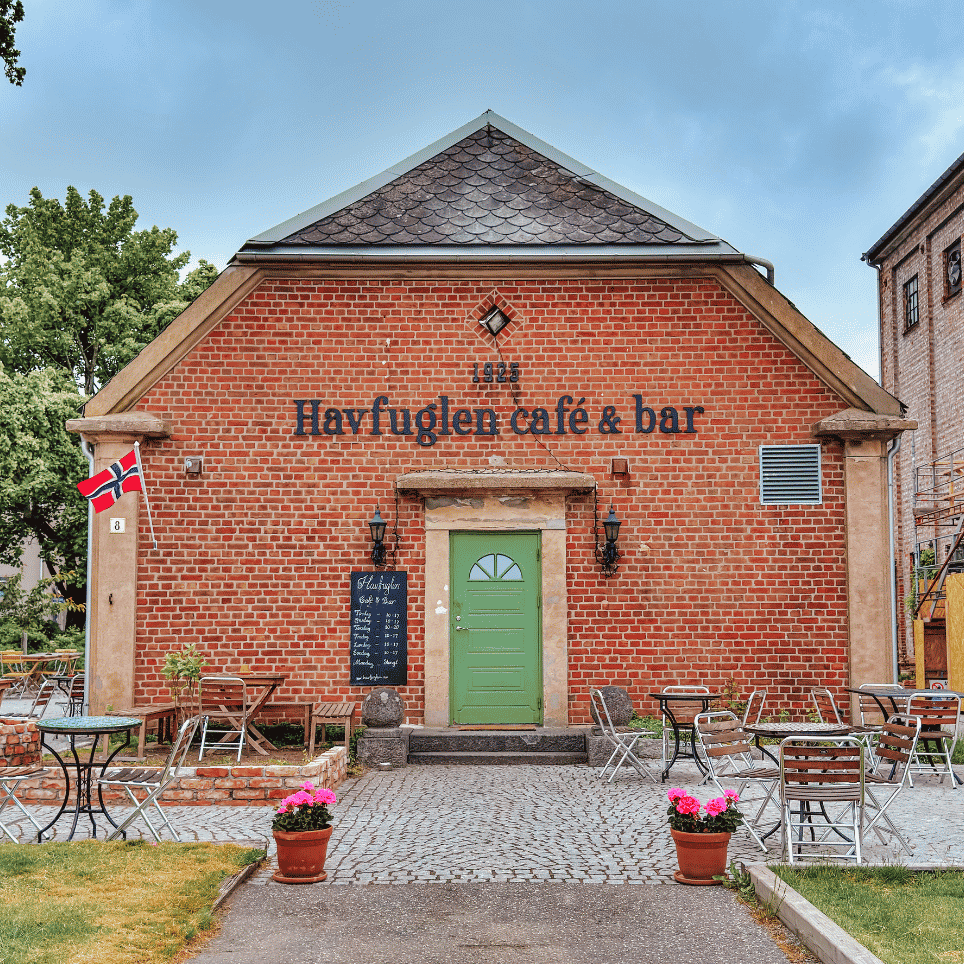 A red timber-framed building dating from 1925, with a sign reading ‘Havfuglen café & bar’, a green front door and outdoor café tables on the cobbled square.