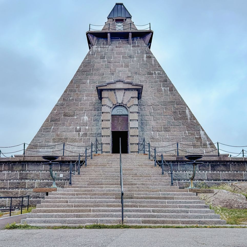 A stone-built, pyramid-shaped monument with steps leading up to a gateway and a watchtower at the top, photographed from below.