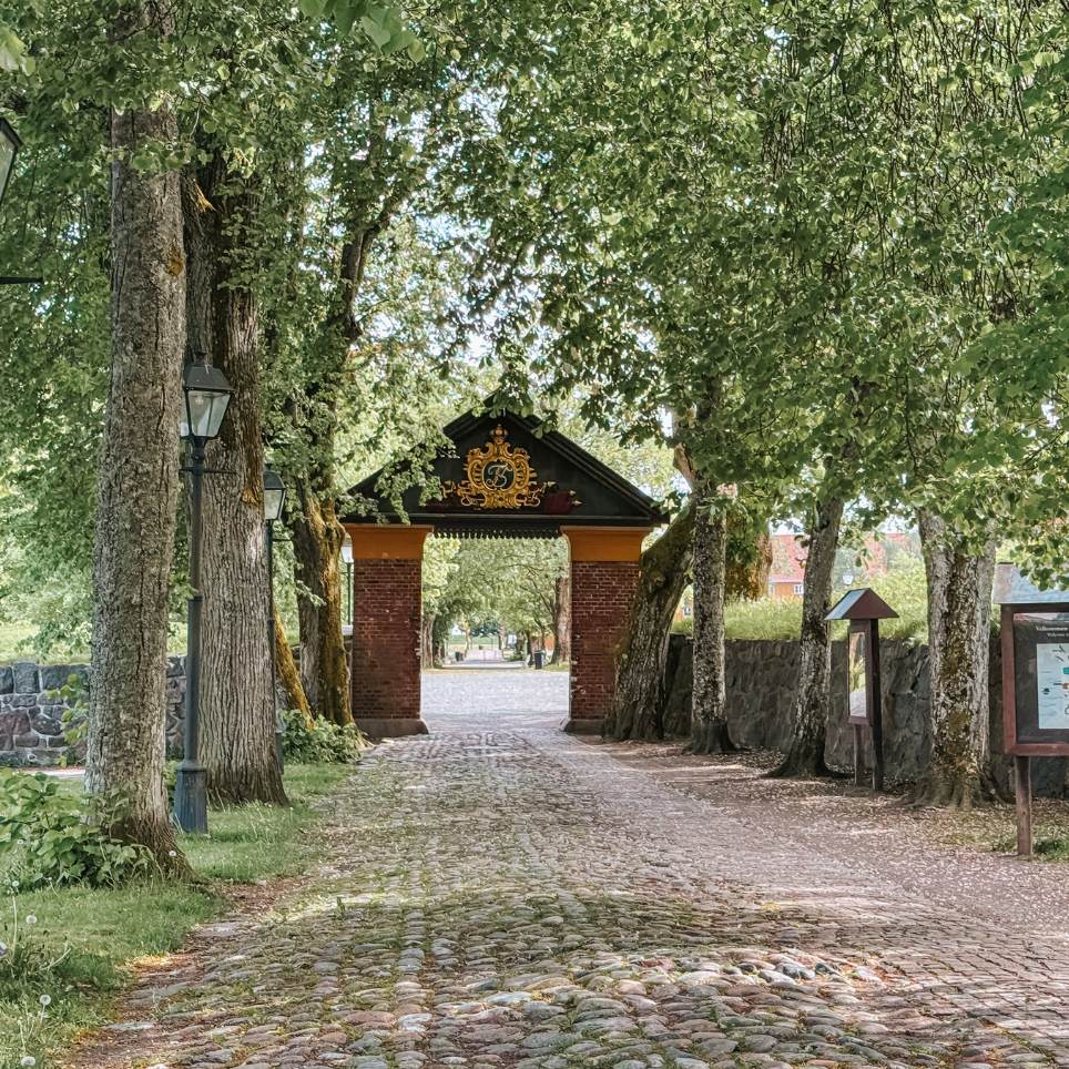 A cobbled avenue lined with large lime trees, leading to an old archway with a gilded monogram above a barrel-vaulted entrance.