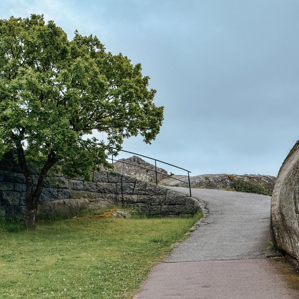 Green deciduous trees by a stone wall and a footpath with a railing that winds its way up a rocky outcrop.